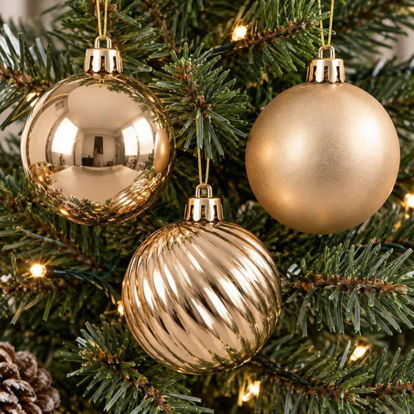 Three gold Christmas tree ornaments hanging on a green pine tree with warm white string lights. One ornament is highly reflective, one is matte, and one has a ribbed textured pattern.