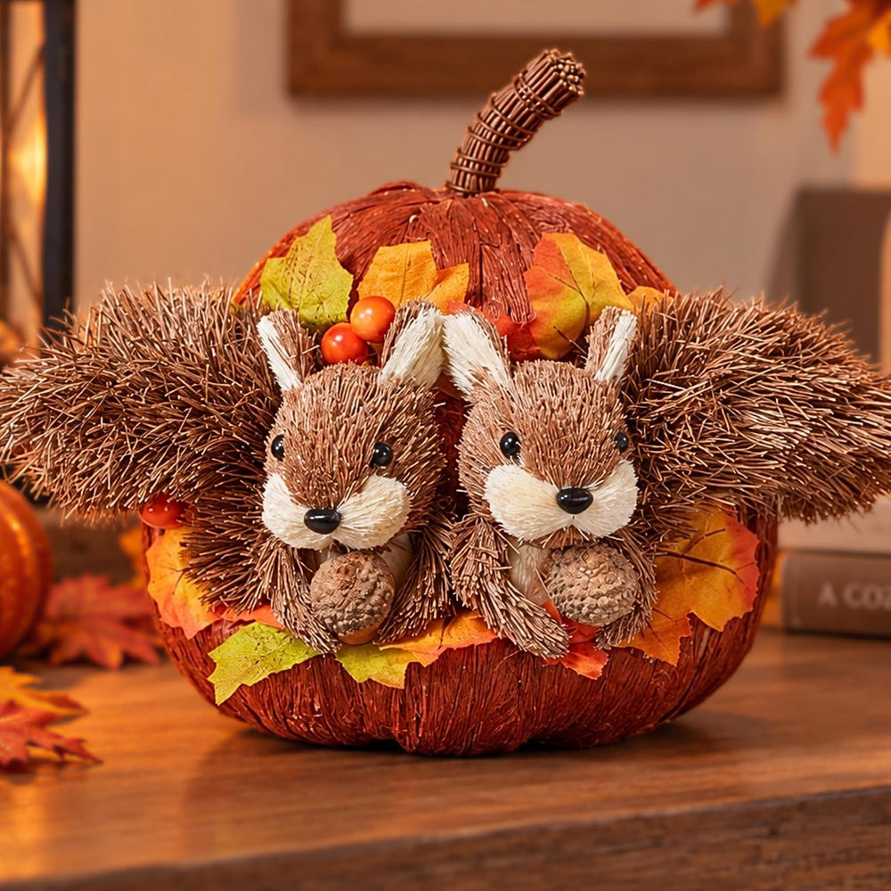 A decorative autumn centerpiece featuring two plush brown squirrel figures nestled inside a wrapped orange pumpkin, surrounded by faux fall leaves and small berries, displayed on a wooden surface.