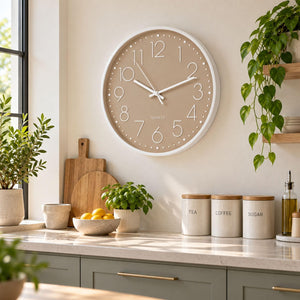 A round wall clock with a beige face and white frame hangs on a kitchen wall above a countertop with canisters, plants, and wooden cutting boards.