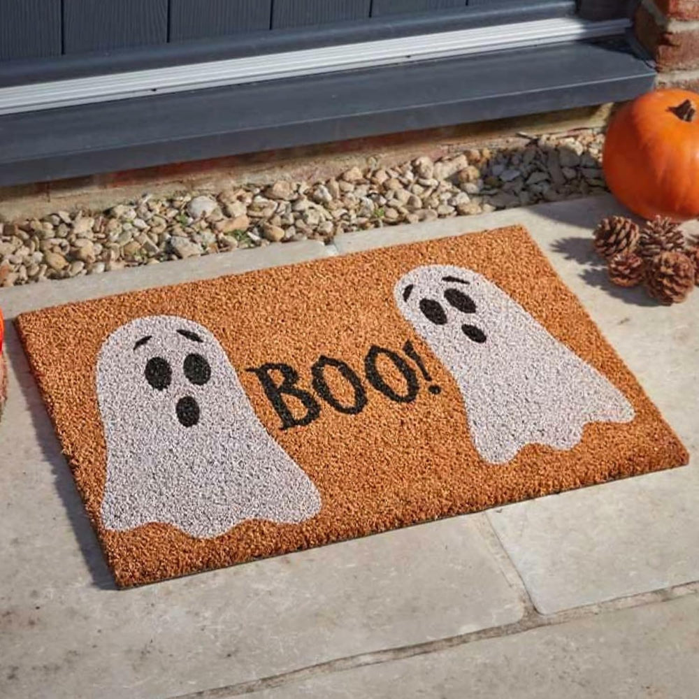 A rectangular brown coir doormat with two white ghost illustrations and the word "BOO!" in black, placed on a stone patio in front of a door with a pumpkin and pinecones nearby.