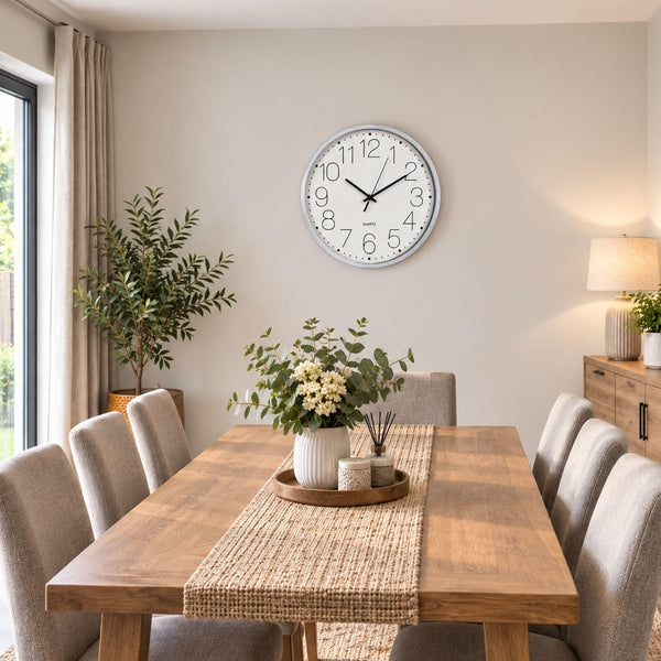A round white wall clock with black numerals and hands hangs above a wooden dining table in a bright, modern dining room with beige upholstered chairs, a woven table runner, and potted plants.