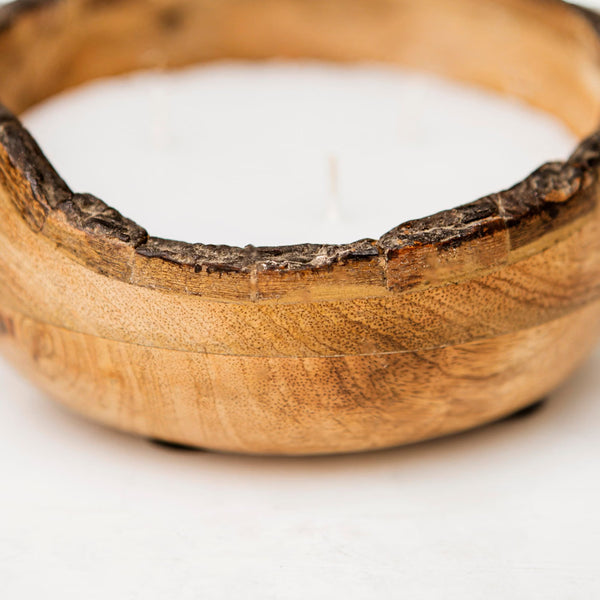 A close-up of a wooden bowl candle holder with a white wax candle inside, showing the natural grain and rough bark edge of the wood.