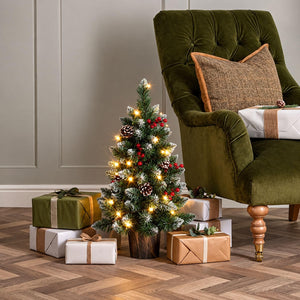 A small frosted Christmas tree with warm white string lights, pinecones, and red berries stands on a wooden floor beside a green velvet armchair, with wrapped gift boxes placed around it in a styled indoor setting.