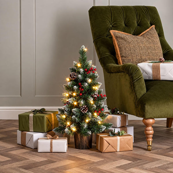 A small frosted Christmas tree with warm white string lights, pinecones, and red berries stands on a wooden floor beside a green velvet armchair, with wrapped gift boxes placed around it in a styled indoor setting.