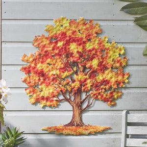 A decorative metal wall art piece shaped like an autumn tree with red, orange, yellow, and green leaves, mounted on a light gray wood siding wall.
