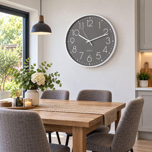 A round wall clock with a gray face and white frame hangs on a light gray wall above a wooden dining table with gray upholstered chairs in a bright, modern kitchen-dining room.