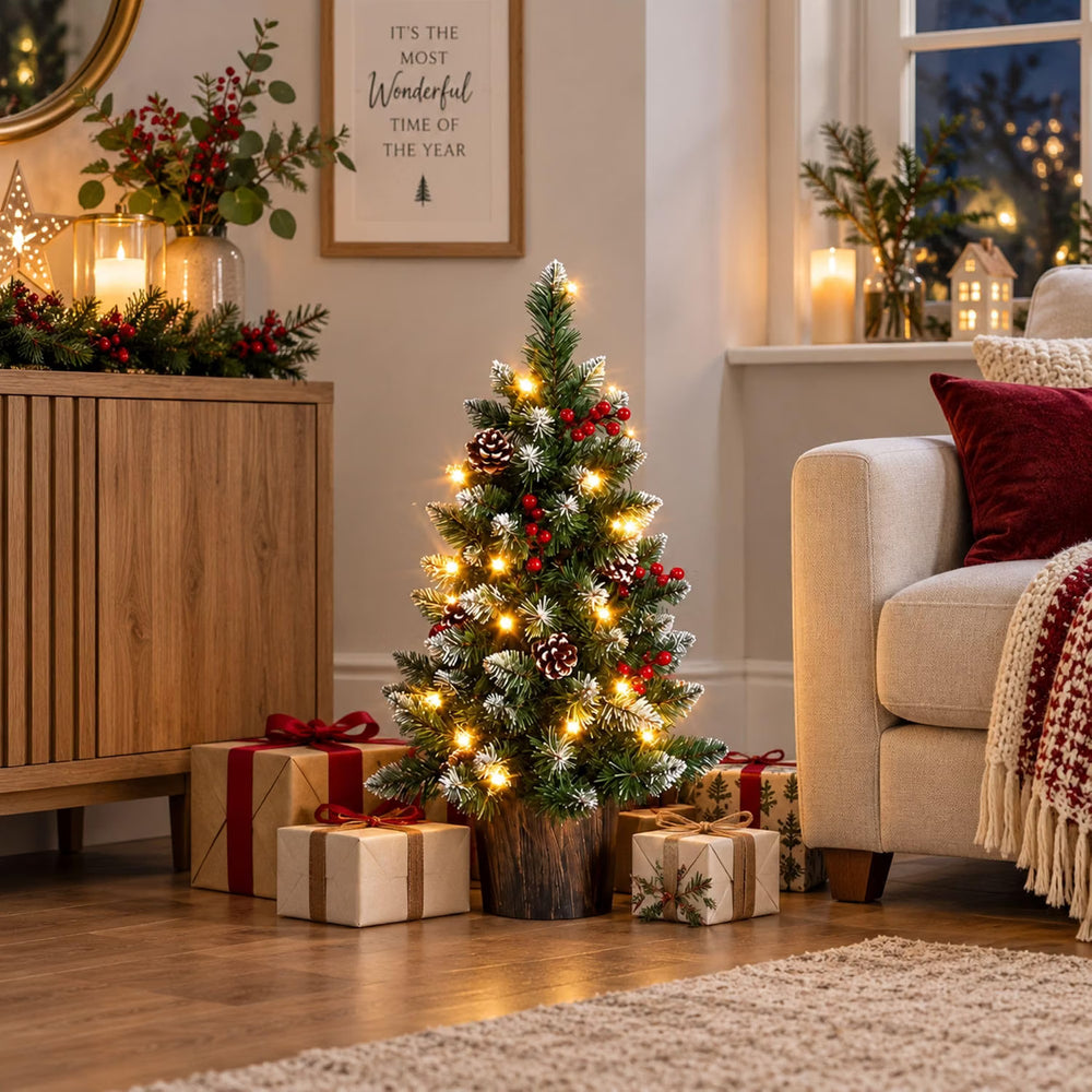 A small artificial Christmas tree with warm white string lights, faux snow, pinecones, and red berries stands on the floor in a cozy living room beside wrapped gifts, a wooden cabinet, and a cream sofa.