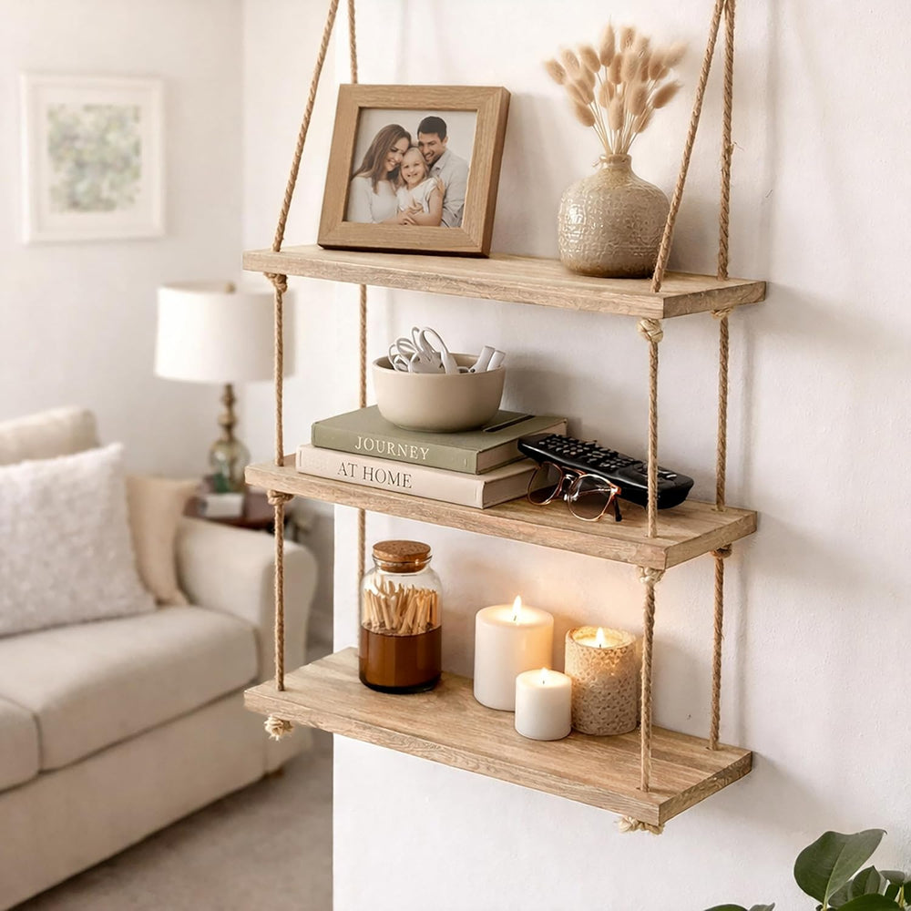 A three-tier floating wooden shelf hanging on a wall in a living room setting, styled with a framed photo, a vase with dried grass, books, a bowl, candles, and a jar.