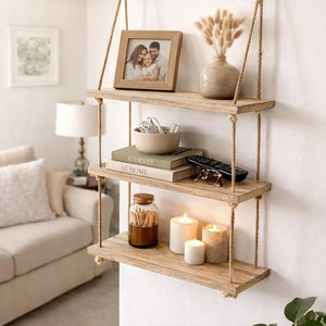 A three-tier floating wooden shelf hanging on a wall in a living room setting, styled with a framed photo, a vase with dried grass, books, a bowl, candles, and a jar.