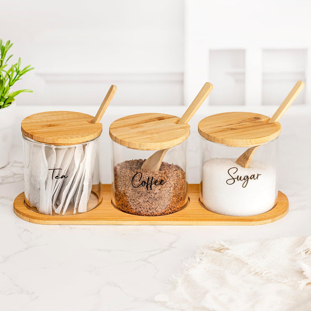 Three clear glass spice jars with bamboo lids and bamboo spoons sit on a matching bamboo tray on a white marble countertop. The jars are labeled Tea, Coffee, and Sugar.
