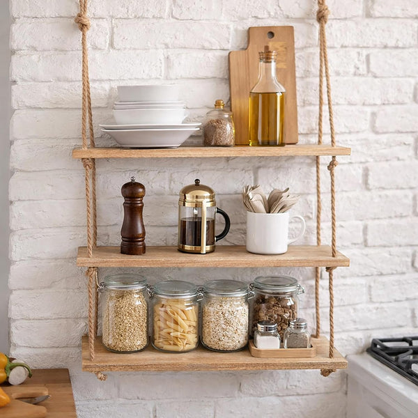 A rustic three-tier floating wooden shelf hanging from rope against a white brick wall, styled with kitchen items including glass jars of grains, a coffee press, a pepper mill, stacked plates, and a bottle of olive oil.