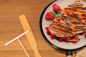 Stack of pancakes with chocolate drizzle and strawberries on a white plate, placed on a wooden table.