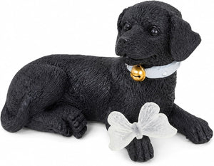 A black dog figurine lying down, wearing a white collar with a gold bell, and holding a white butterfly ornament, photographed on a plain white background.