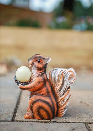 A decorative resin squirrel figurine holding a white acorn, standing on a stone patio with a blurred outdoor garden background.