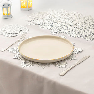 A round off-white ceramic plate with a thin gold rim, styled on a white tablecloth with white paper snowflakes, silver cutlery, and decorative lanterns.
