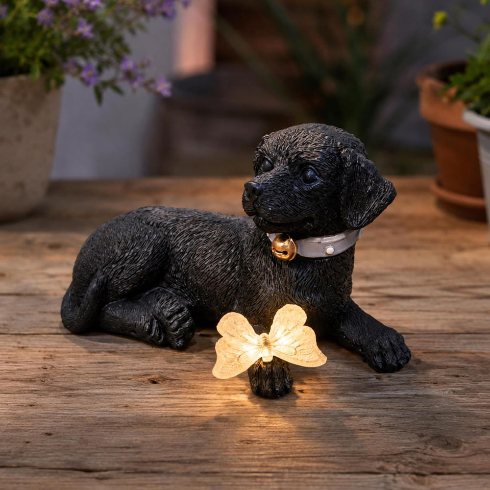 A black dog-shaped garden statue lying down, wearing a white collar with a gold bell, placed on a wooden surface next to a small glowing butterfly light. Potted plants are blurred in the background.