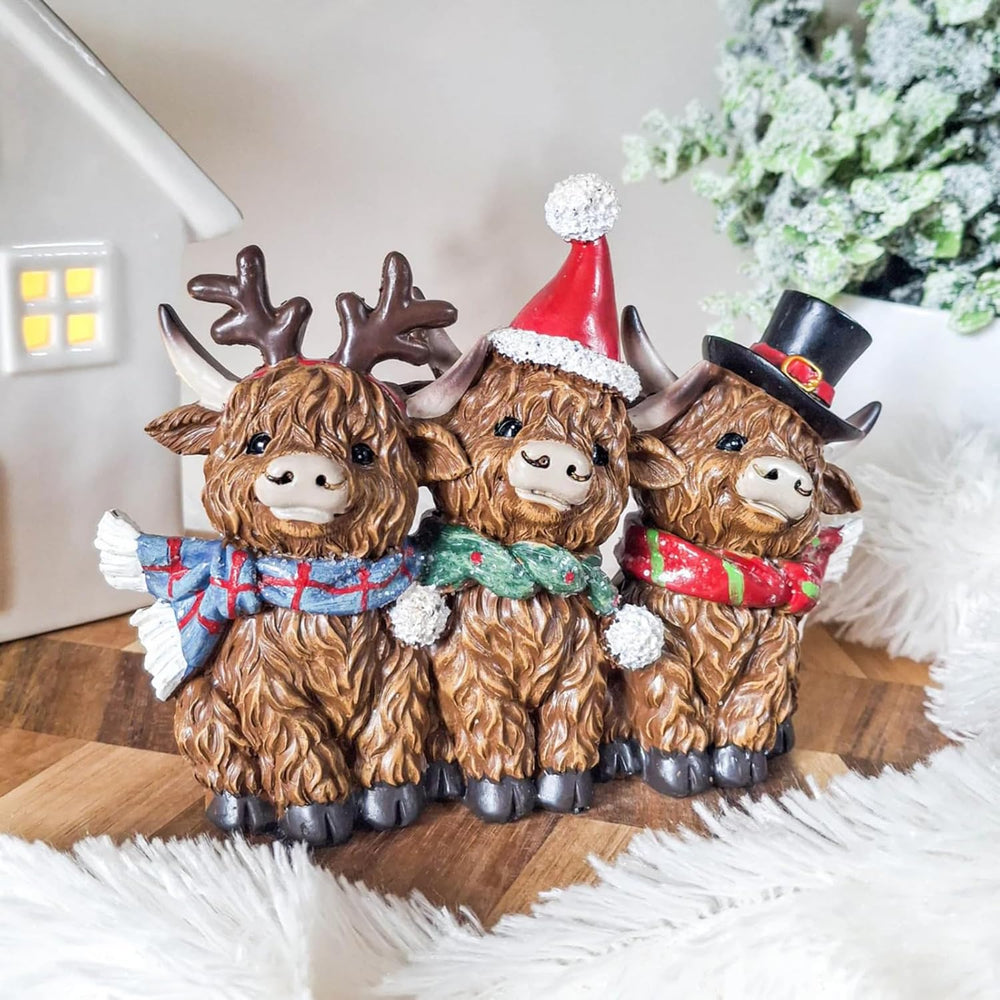 Three decorative Highland cow figurines wearing Christmas hats and scarves on a wooden surface.