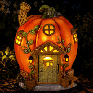 A decorative pumpkin-shaped house night light with a glowing warm yellow interior, detailed door, windows, vine accents, and small lanterns, shown outdoors against a dark foliage background.