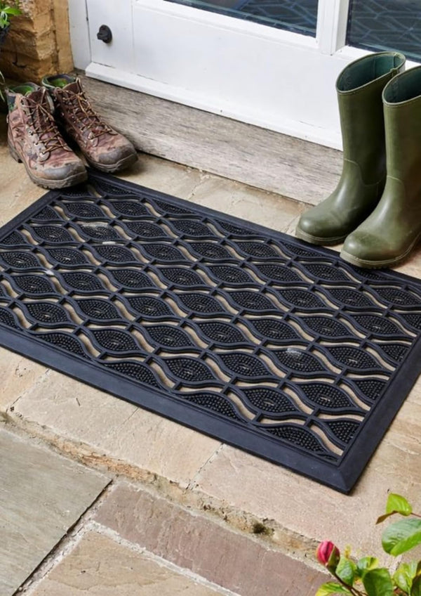 Black doormat with textured pattern on a wooden floor, next to green boots and a white door.
