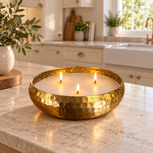 A hammered gold metal candle holder containing three lit white candles, placed on a light beige marble kitchen countertop with a blurred kitchen background.
