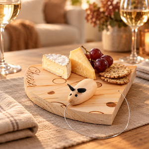 A wooden cheese board with engraved cutouts and the word "Cheese" on the side, styled on a table with wine glasses, crackers, grapes, and a small wooden mouse-shaped cheese pick. The background shows a softly blurred living room setting.