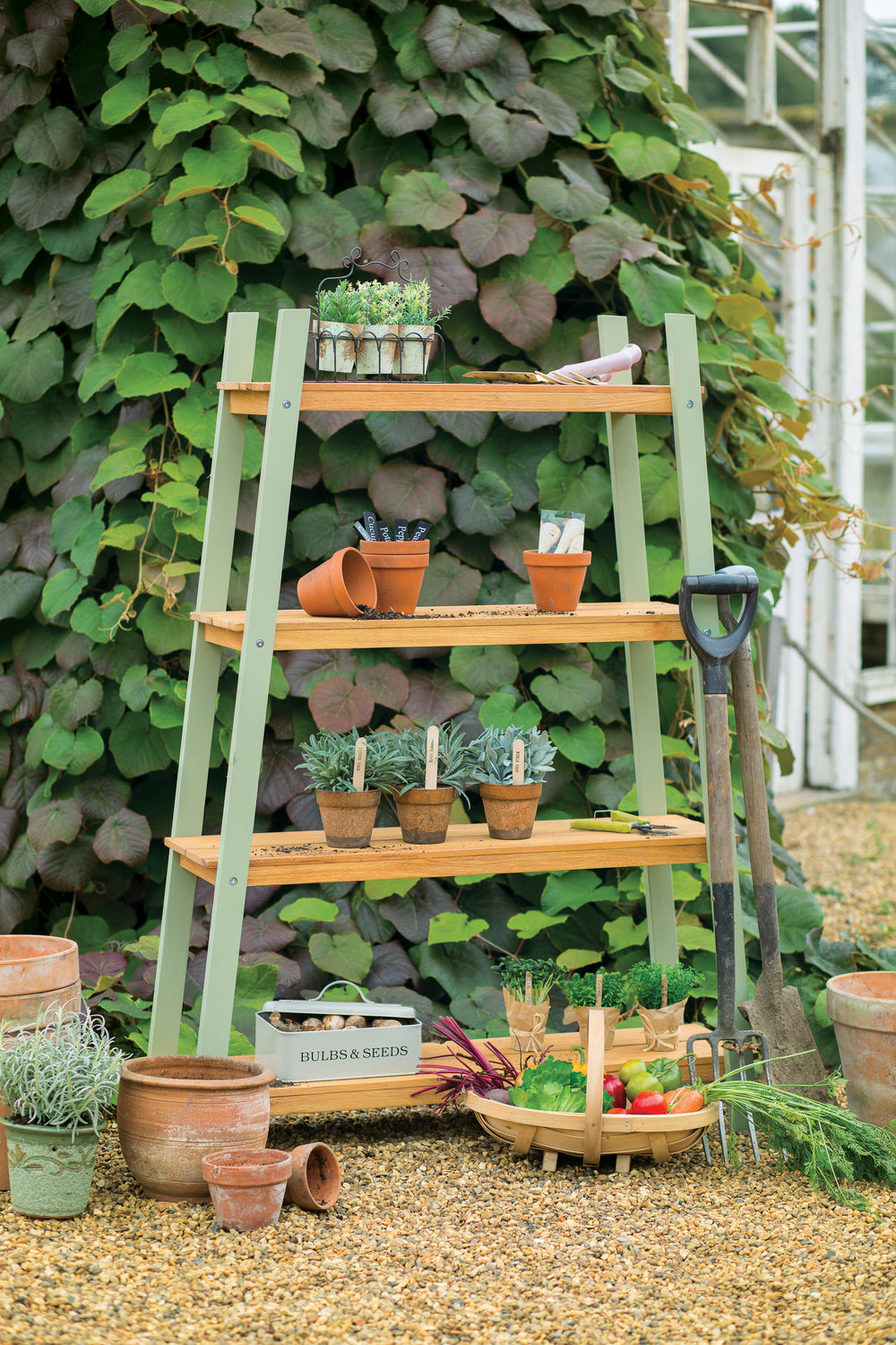 A green wooden plant shelf with four tiers, displaying various plants and pots, placed outdoors possibly in a garden setting.