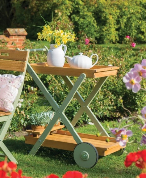 A green wooden drinks trolley with wheels, featuring a tray on top with a white teapot and flowers, set in a garden environment.
