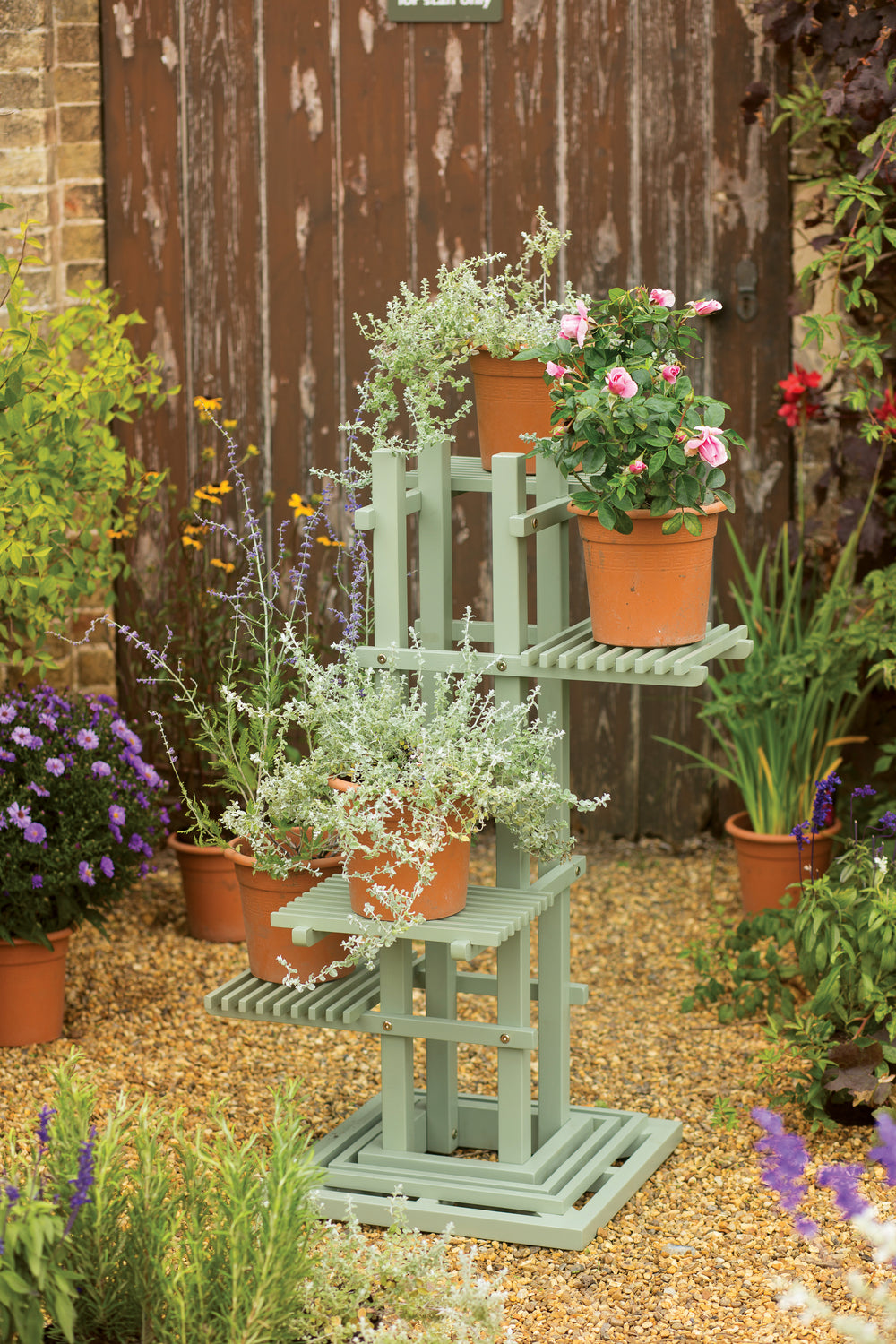 A green, multi-tiered wooden plant stand displaying various potted plants, placed outdoors near a wooden fence and colorful flowers.