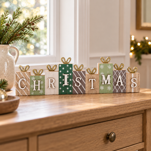 A decorative wooden block sign spelling CHRISTMAS arranged on a wooden cabinet in a real indoor setting with a window, holiday greenery, and warm string lights in the background.