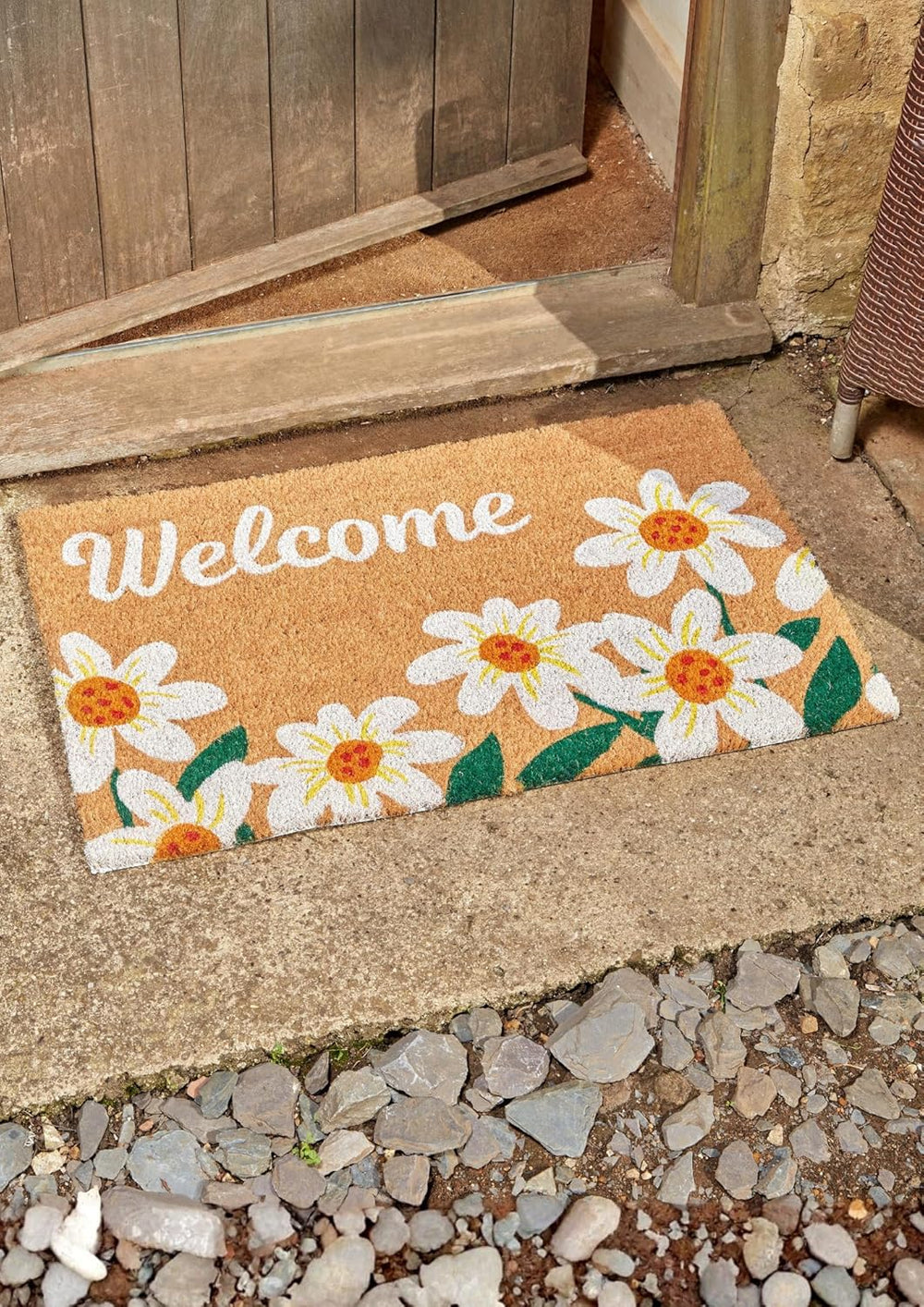 Doormat with daisy design and 'Welcome' text on a stone pathway.