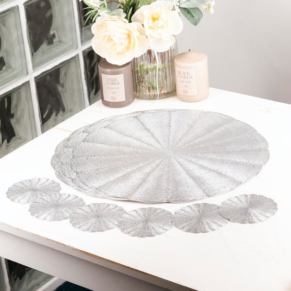 A stack of round silver glitter placemats with a scalloped edge is displayed on a white table, with several placemats laid out in a row in front. Decorative props including a vase of cream roses, a candle, and a jar are placed behind the product.