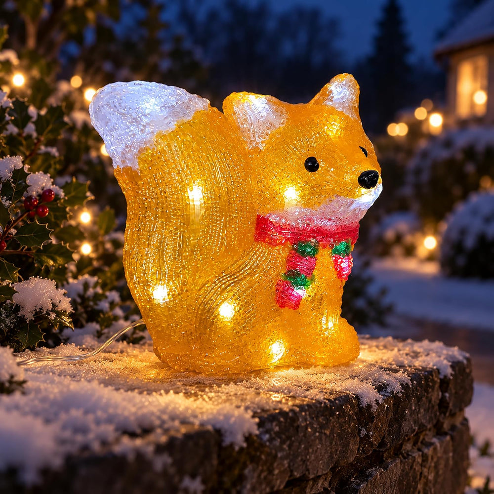 A glittery golden fox-shaped outdoor light with built-in white LED lights, wearing a red and green striped scarf, displayed on a stone surface at dusk.