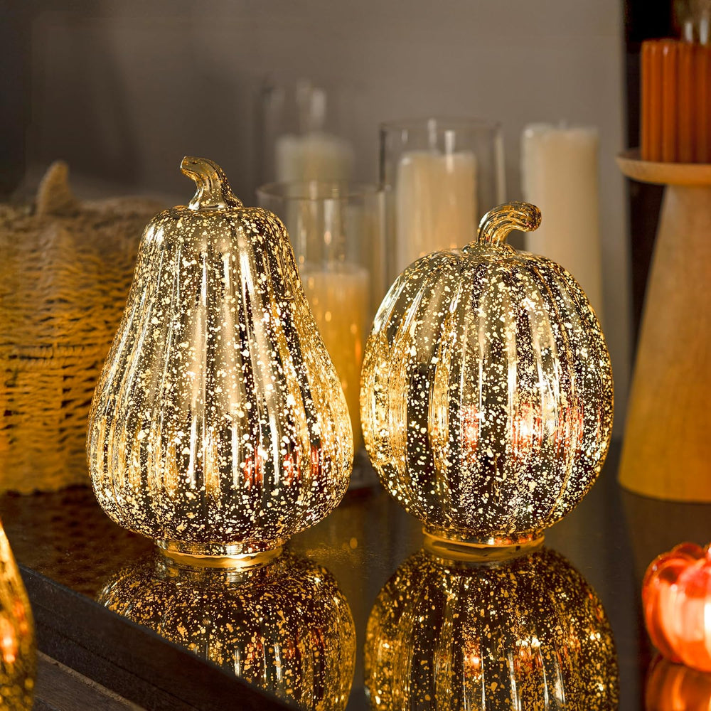 Two glittery pumpkin-shaped lamps on a reflective surface with candles and a basket in the background.