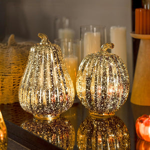 Two glittery pumpkin-shaped lamps on a reflective surface with candles and a basket in the background.