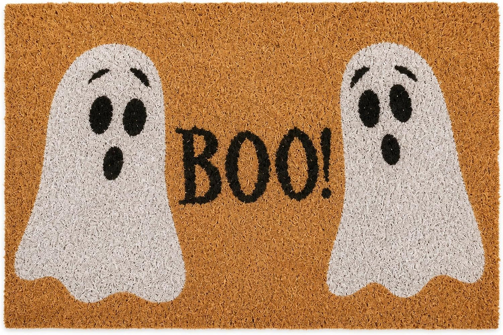 A rectangular brown coir doormat with two white ghost illustrations and the word "BOO!" in black, placed on a stone patio in front of a door with a pumpkin and pinecones nearby.