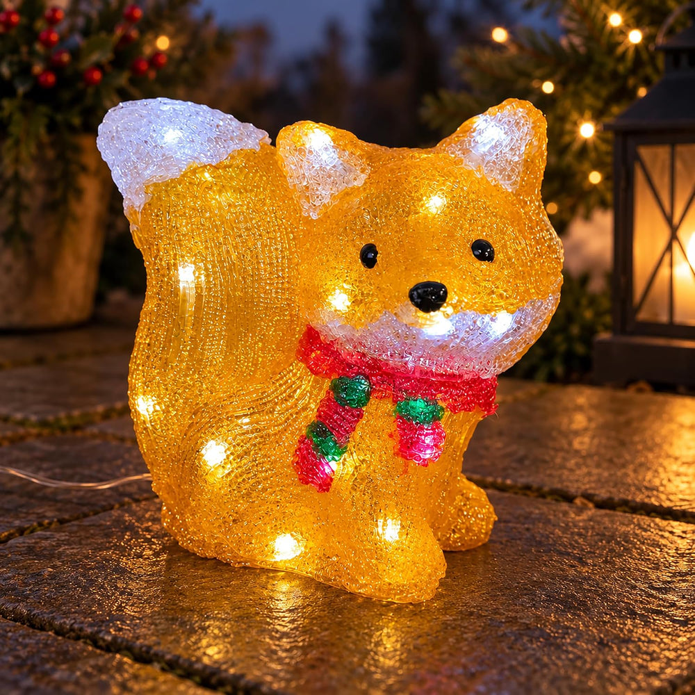 A glittery golden fox-shaped outdoor light with built-in white LED lights, wearing a red and green striped scarf, displayed on a stone surface at dusk.
