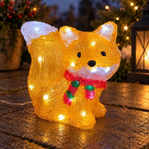 A glittery golden fox-shaped outdoor light with built-in white LED lights, wearing a red and green striped scarf, displayed on a stone surface at dusk.