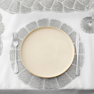 A place setting featuring a round cream-colored dinner plate with a gold rim, layered on top of a textured silver scalloped charger plate, set on a white tablecloth with a fork and knife.