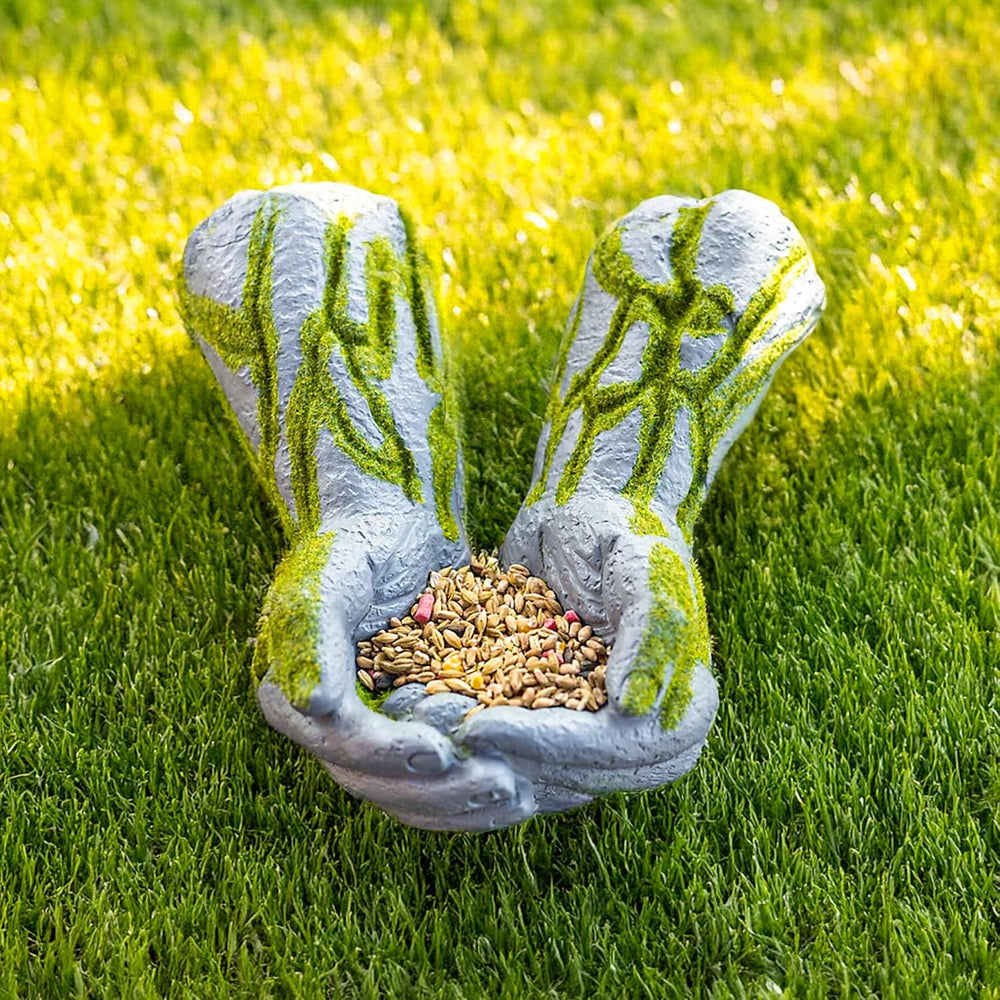 A garden-style hand-shaped planter or seed holder made of textured gray material with green mossy detailing, holding a pile of birdseed on a bright green grass lawn.