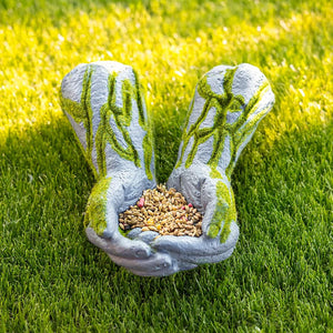 A garden-style hand-shaped planter or seed holder made of textured gray material with green mossy detailing, holding a pile of birdseed on a bright green grass lawn.