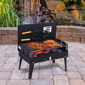 A black metal folding picnic grill with legs, shown open with skewers of food cooking over visible coals. The grill is placed on a stone patio in front of a landscaped rock wall with a small waterfall and greenery.