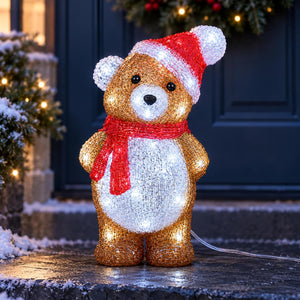 A glittery teddy bear Christmas light decoration wearing a red and white Santa hat and red scarf, standing on a snowy doorstep in front of a dark blue door.