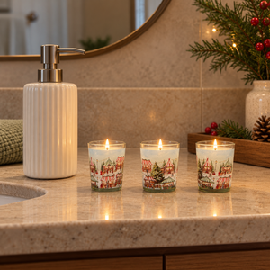 Three lit holiday-themed glass jar candles with a red and white village print sit on a beige stone bathroom counter next to a ribbed white soap dispenser.