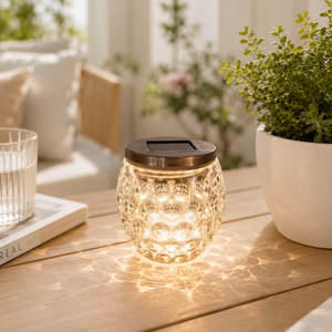 Solar-powered glass lantern on a wooden table with plants and a glass in the background