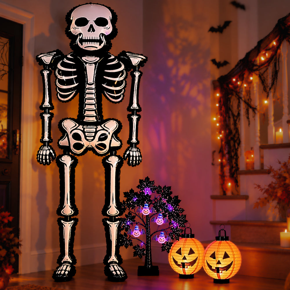 A large Halloween skeleton decoration stands in a decorated home hallway with warm string lights, jack-o'-lantern lanterns, and a small spooky tree.