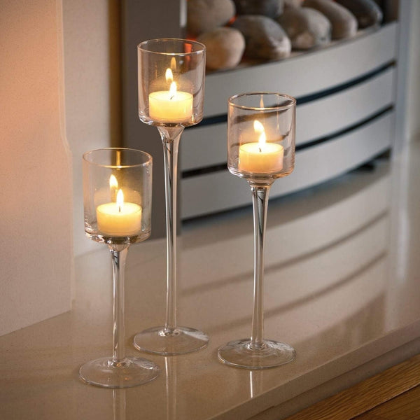 Three clear glass candle holders with long stems, each containing a lit tealight candle, arranged on a glossy tabletop in an indoor setting.