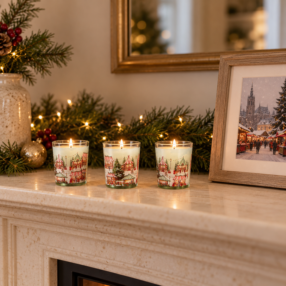 Three lit holiday-themed glass candles with red and green winter village illustrations are arranged on a cream stone mantel, surrounded by pine garlands, warm string lights, a decorative vase, and a framed winter scene.