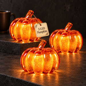 Three glowing orange LED pumpkin lights with a crackle glass texture, displayed on a dark stone surface with a hanging tag that reads '3 LED Pumpkins'.