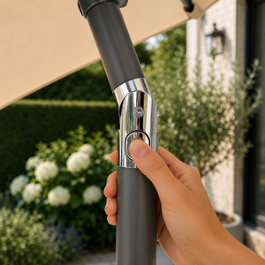 A hand holding a dark gray patio umbrella pole with a chrome button, set against a blurred outdoor garden background.