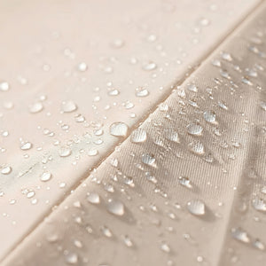 Close-up of a light beige fabric surface with clear water droplets beaded on top and sliding off a slightly textured section, demonstrating a water-repellent finish.